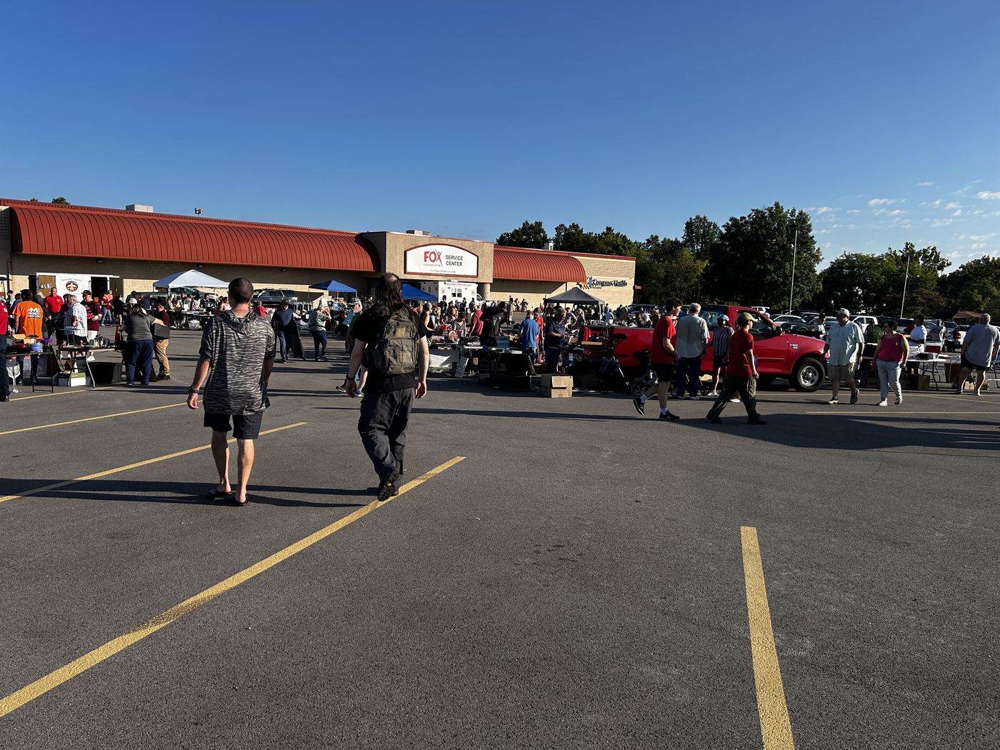 Shoppers crossing the Fox Service Center parking lot toward vendor tents and tables lined up along the front of the building in the morning sun