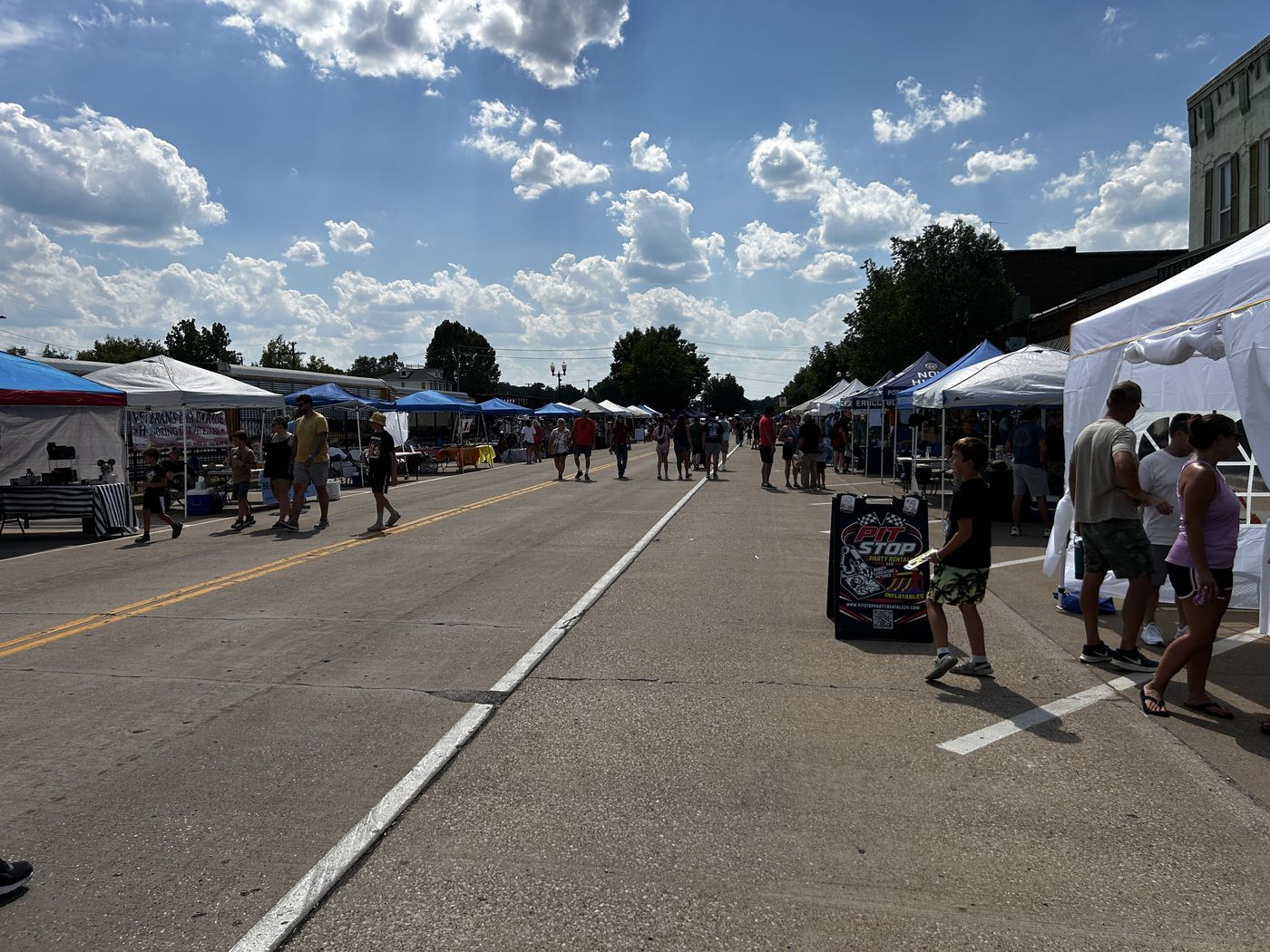 Main Street in De Soto closed to traffic, with white and blue vendor tents down both sides and shoppers walking between them under a blue sky