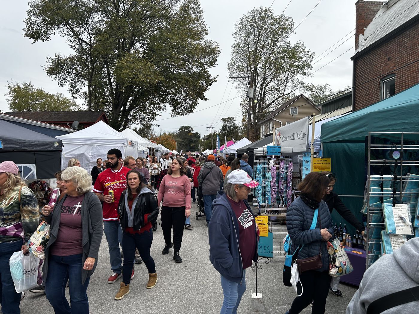 Shoppers walking the closed street between rows of white vendor tents, with a brick storefront and trees behind