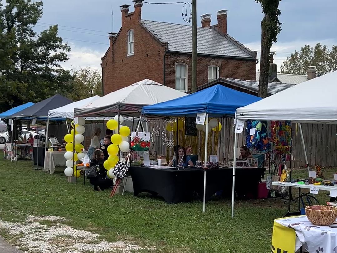 Canopy tents in a row on the grass, yellow and white balloons at one booth and tables of handmade goods, with an old red brick house behind