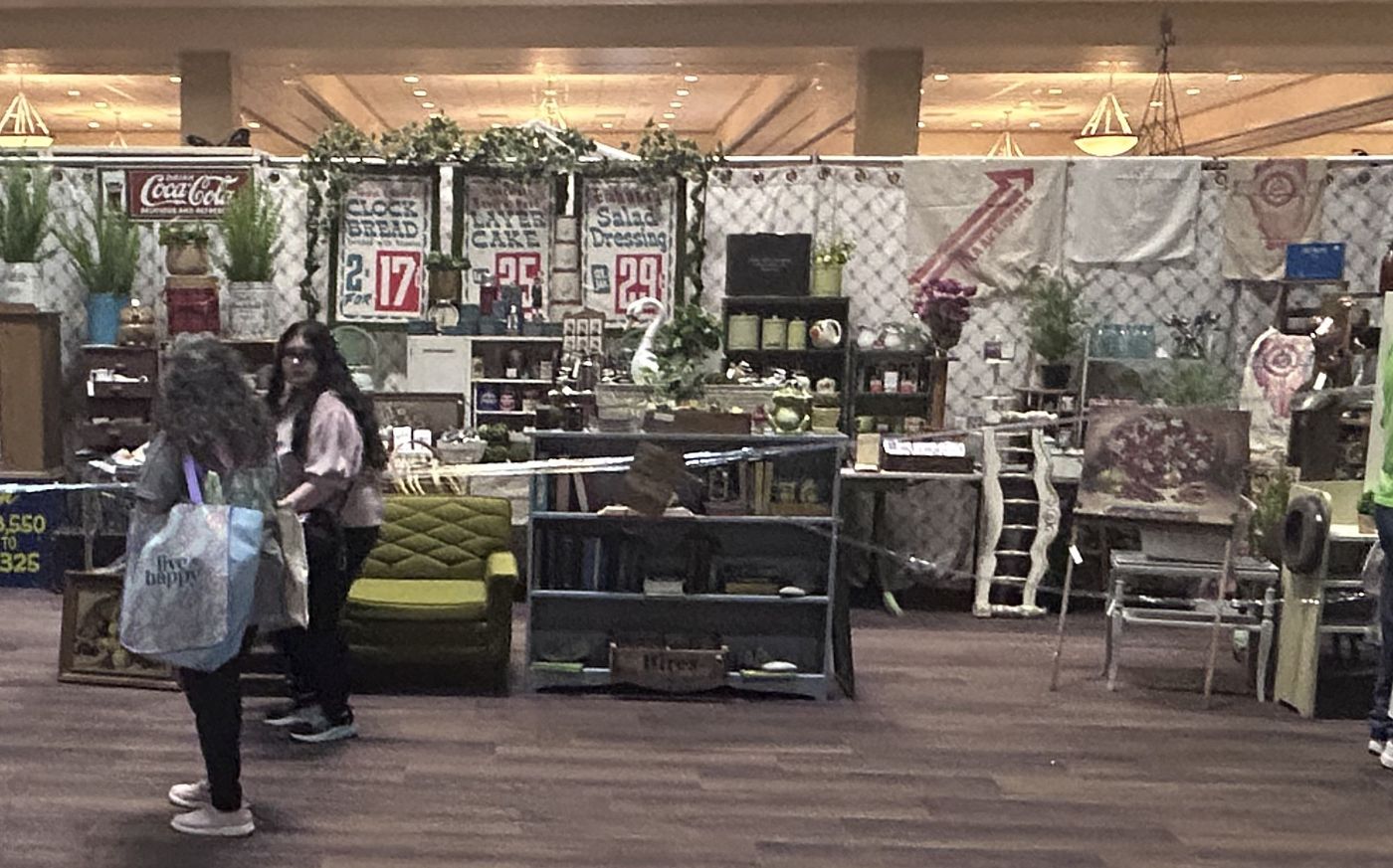 Shoppers with tote bags in the aisle beside a vintage dealer stall hung with old enamel advertising signs, under the hall chandeliers